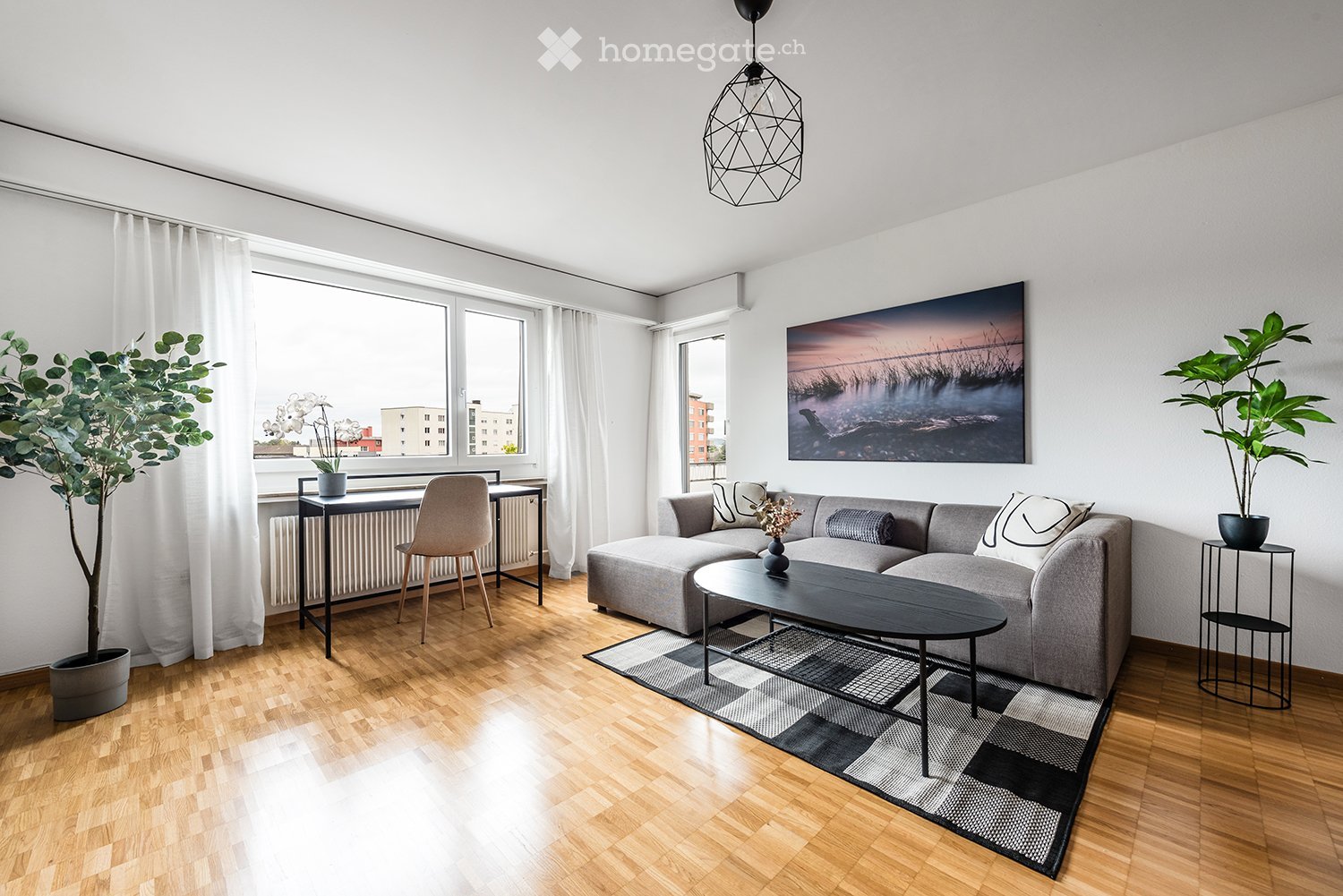 Modern living room with grey sectional sofa, black coffee table, wooden floor, large windows with white curtains, and decorative plants.
