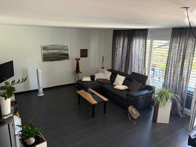 Modern living room with dark tiled flooring, large black sectional sofa, wooden coffee table, floor lamp, television, and potted plants. Large windows with grey curtains lead to a balcony.