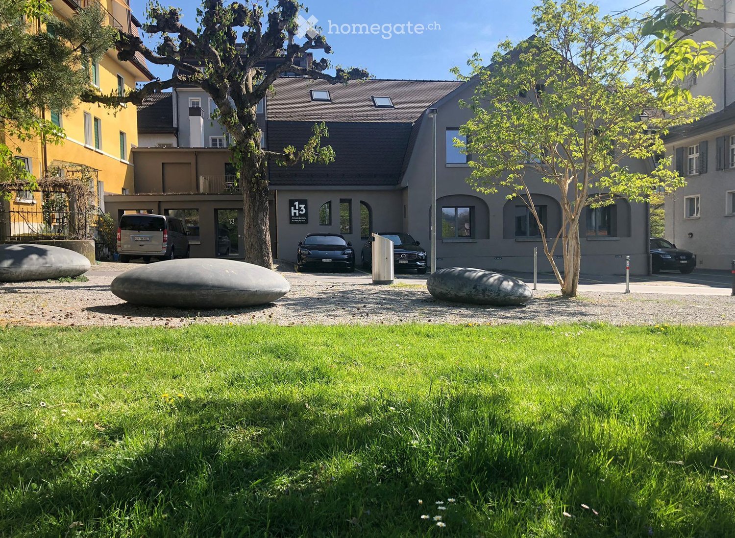 Well-maintained garden with green grass, a gravel driveway, and a modern building with a grey facade and large windows. Several cars are parked in front of the building, and there are two large, sculptural stone forms in the garden area.