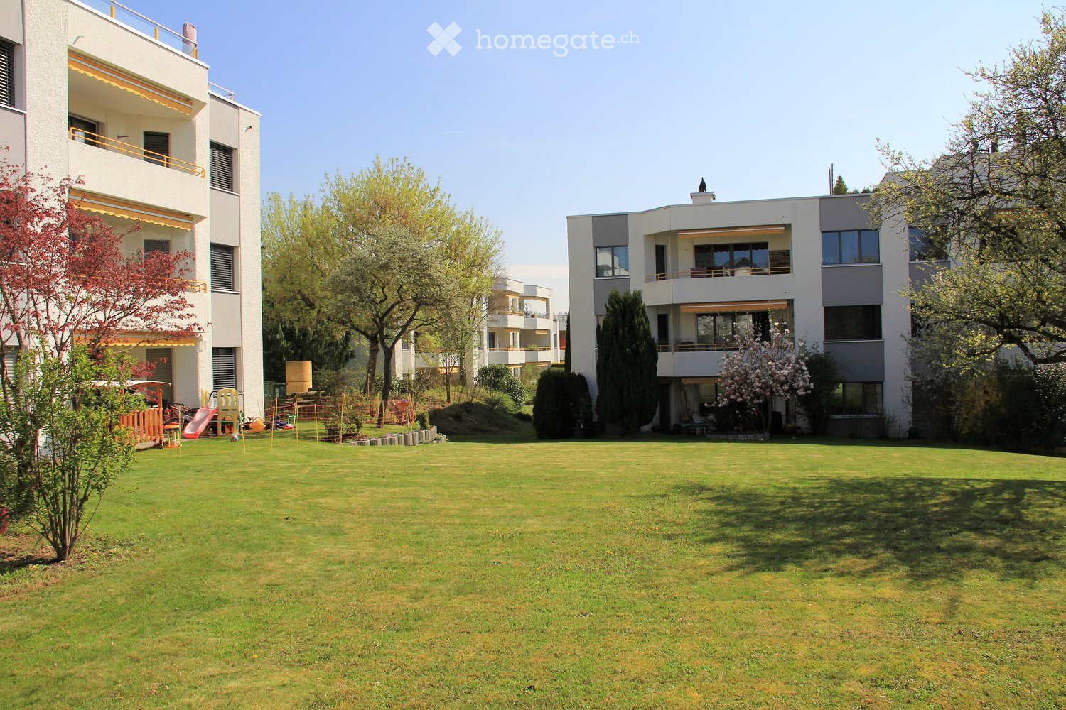 Well-maintained lawn with modern apartment buildings in the background, featuring balconies, trees, and a small play area with a slide and chairs.