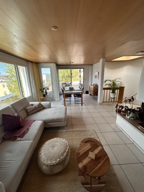 Bright living and dining area with large windows, wooden ceiling, tiled floor, white sectional sofa, ottoman, coffee table, dining table with chairs, side table, and plants.