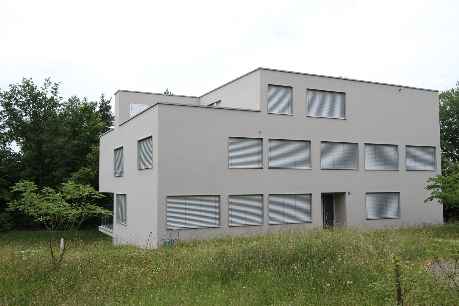 Modern two-story house with grey exterior, multiple windows with blinds, and a small balcony on the left side.