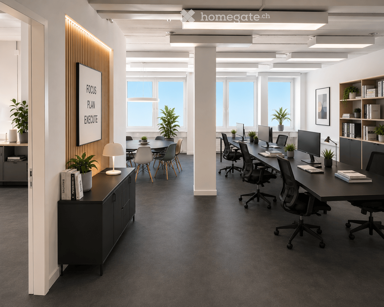 Modern office space with desks, chairs, multiple computer screens, keyboards, a round meeting table, potted plants, and decorative lamps.