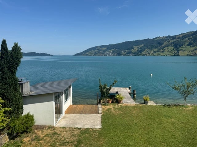 A modern house with a view of a lake and mountains, featuring a wooden deck, potted plants, and a well-maintained lawn.