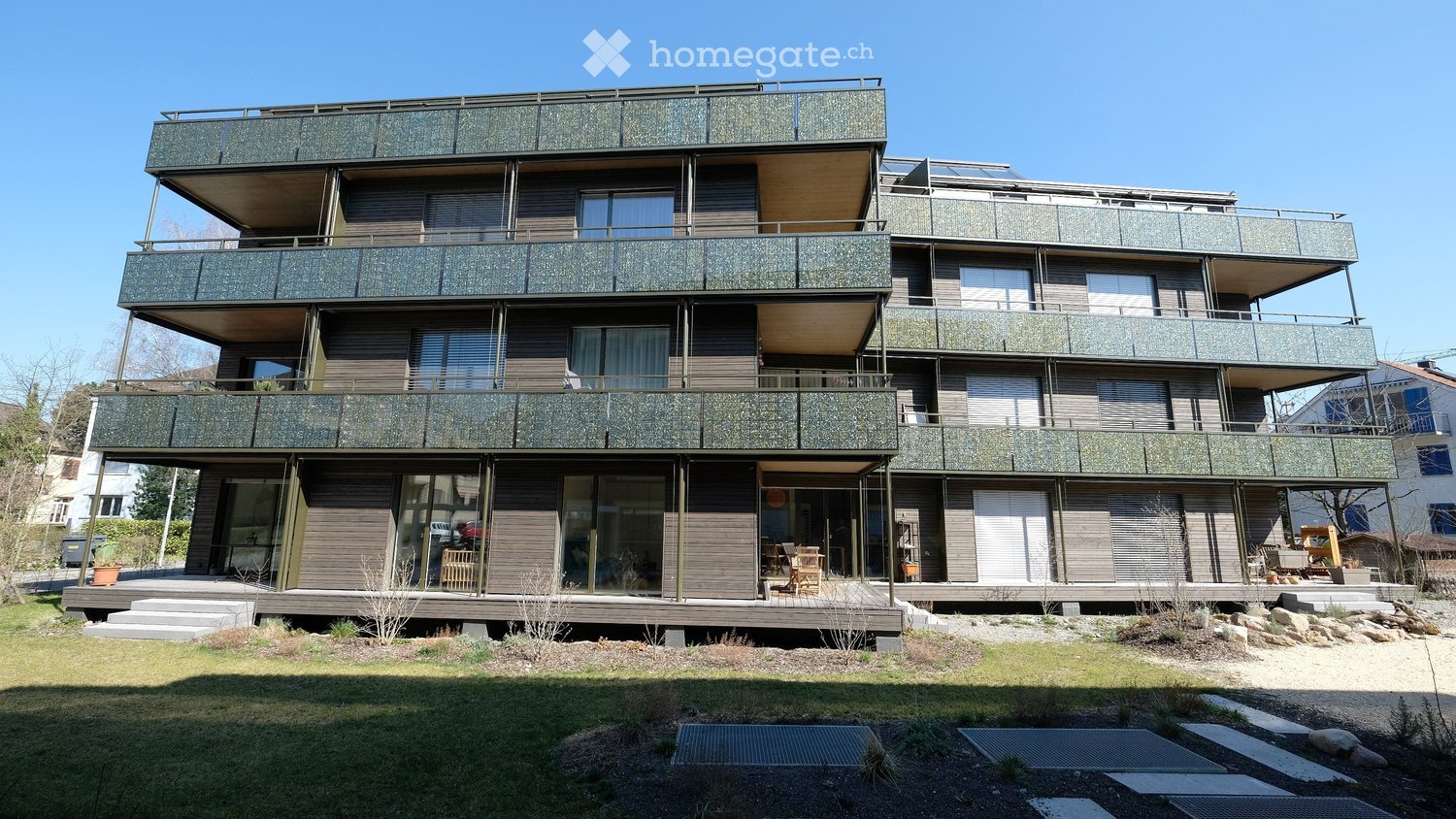 Modern multi-story building with green balconies and large windows, surrounded by a landscaped garden area with stepping stones and rocks.