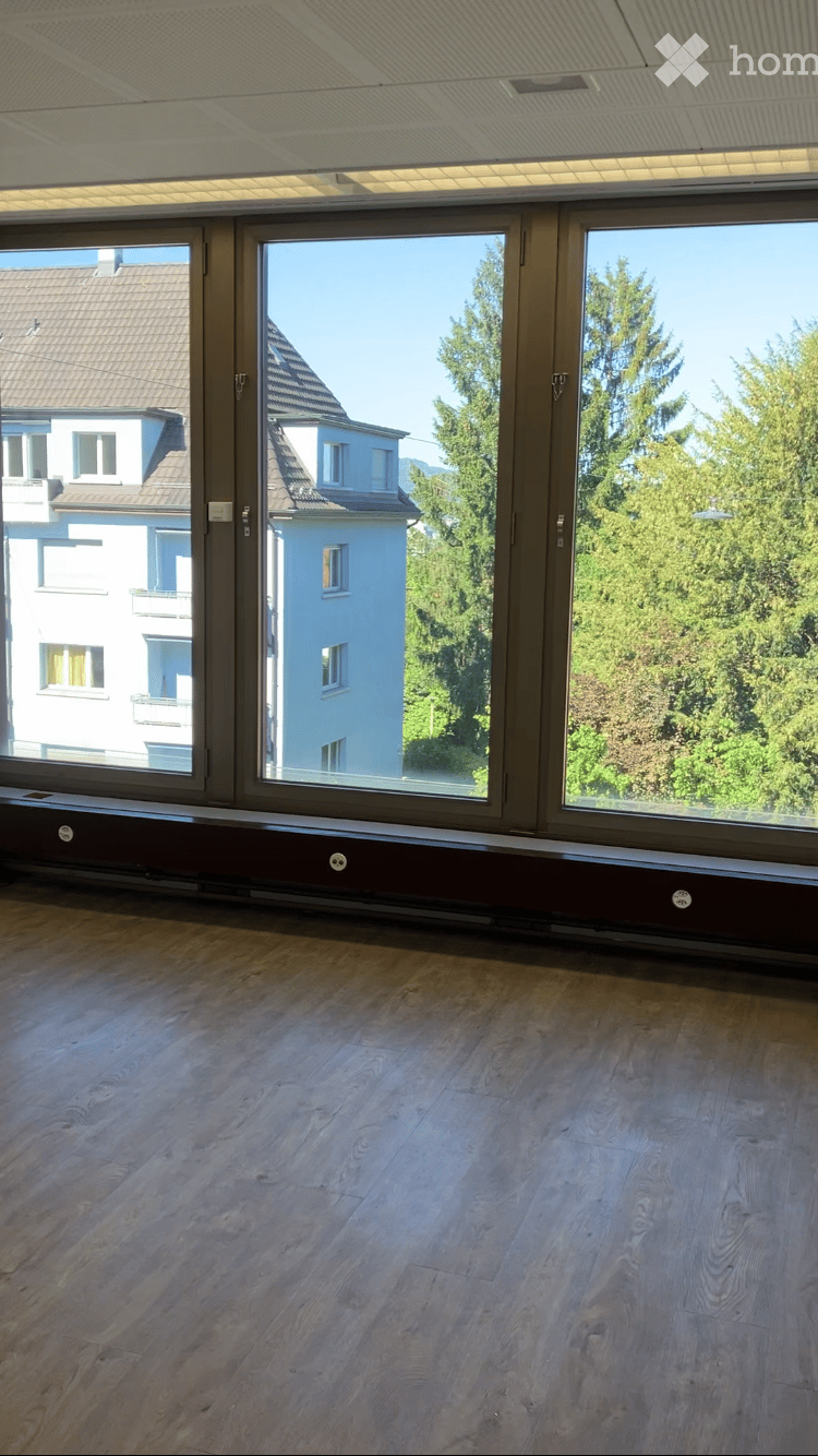Empty room with wooden flooring and large windows offering a view of neighboring buildings and greenery.