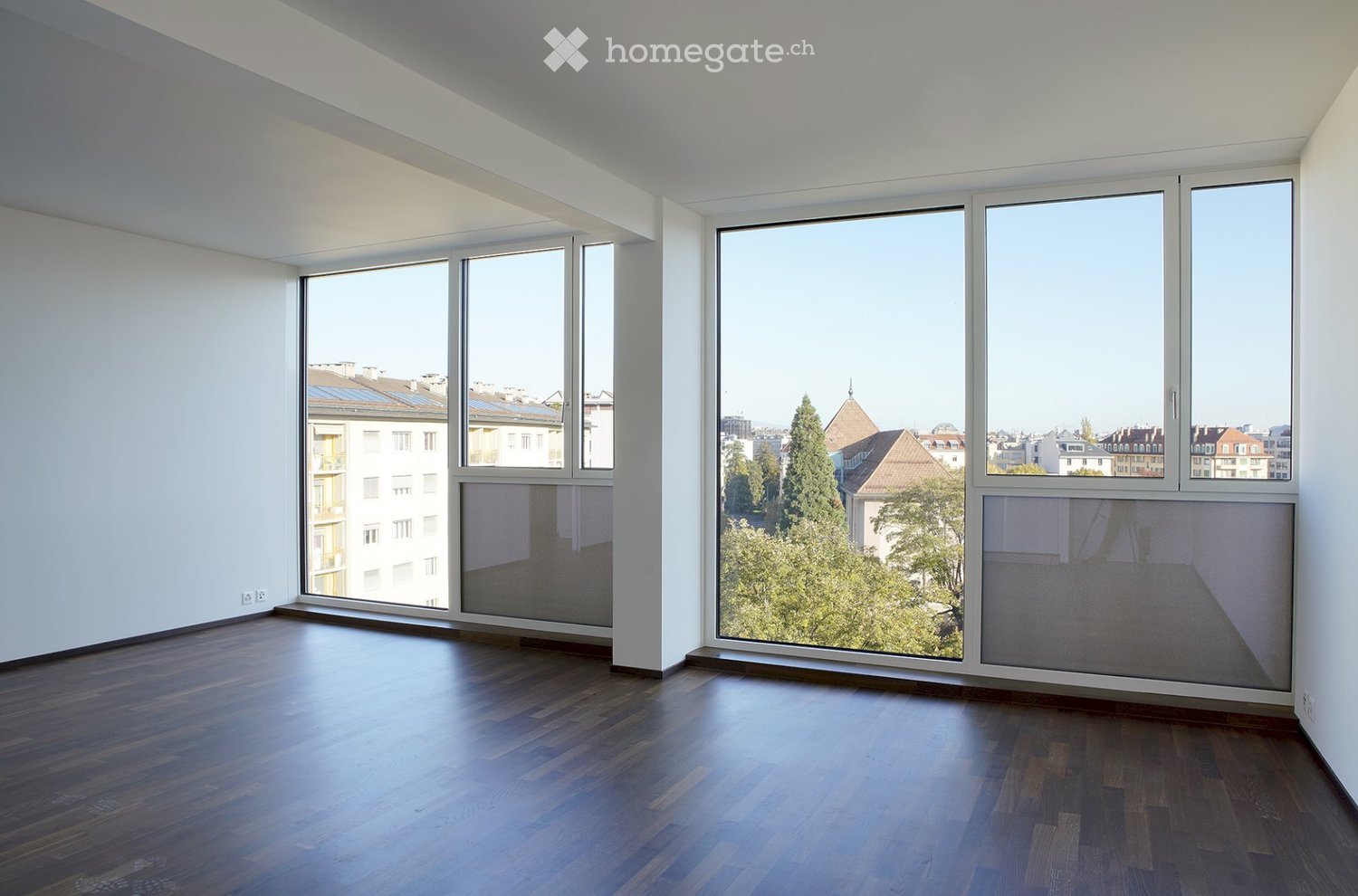 Empty room with wooden flooring and large windows offering a cityscape view. The walls are white, and the space is well-lit with natural light.