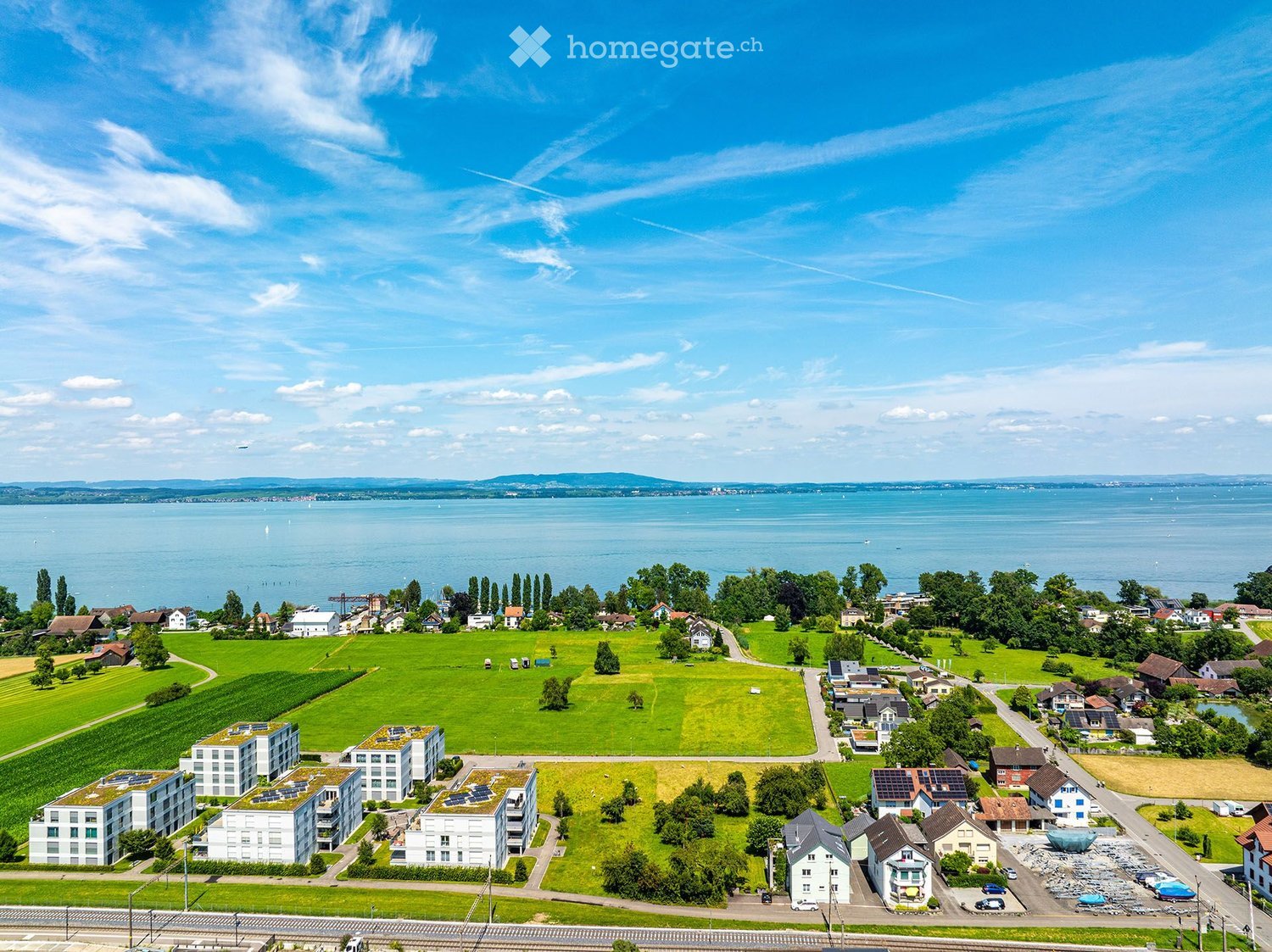Aerial view of a suburban area with residential buildings, green fields, and a large body of water in the background.
