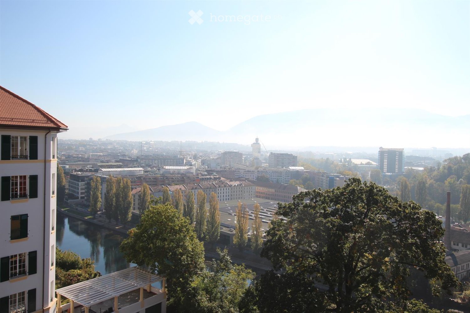 A panoramic view of a cityscape with a river running through it, surrounded by buildings, trees, and distant mountains.