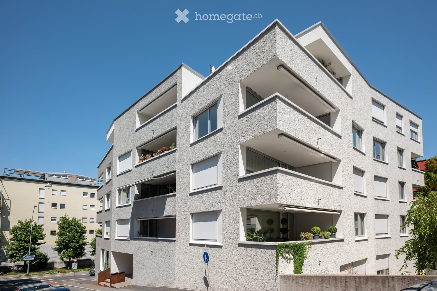 Modern multi-story residential building with white facade, large windows, and balconies adorned with plants and flowers. The building features a clean, contemporary design with geometric shapes and a raised ground floor entrance.