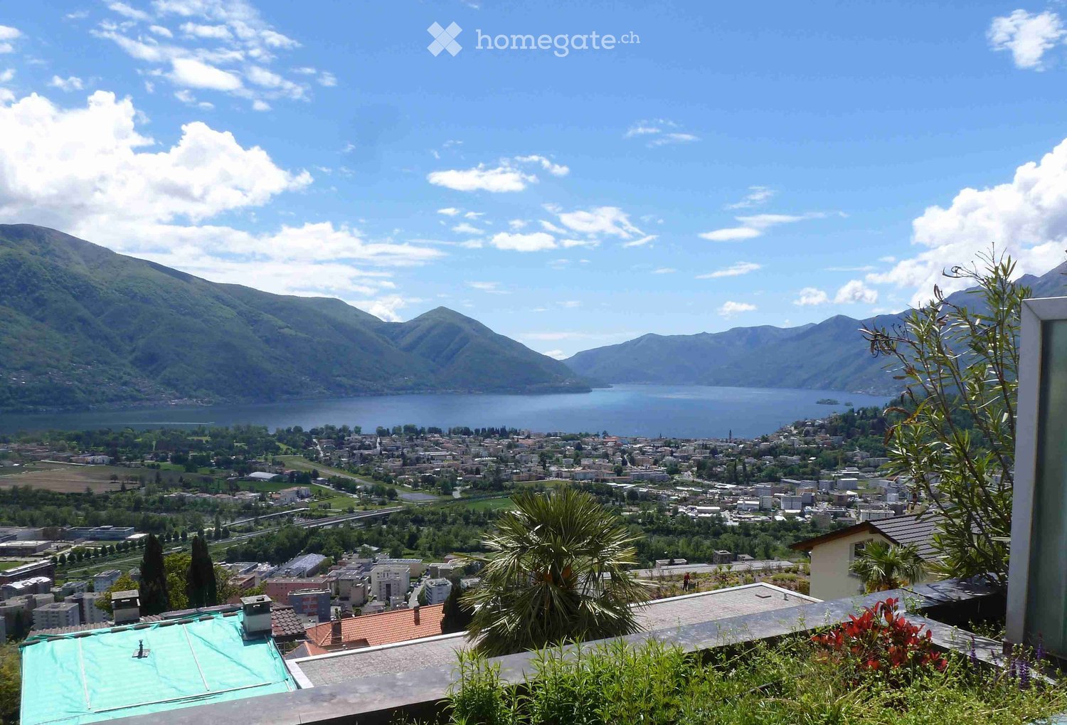 Panoramic view from a balcony or terrace overlooking a town, lake, and surrounding mountains under a partly cloudy sky.