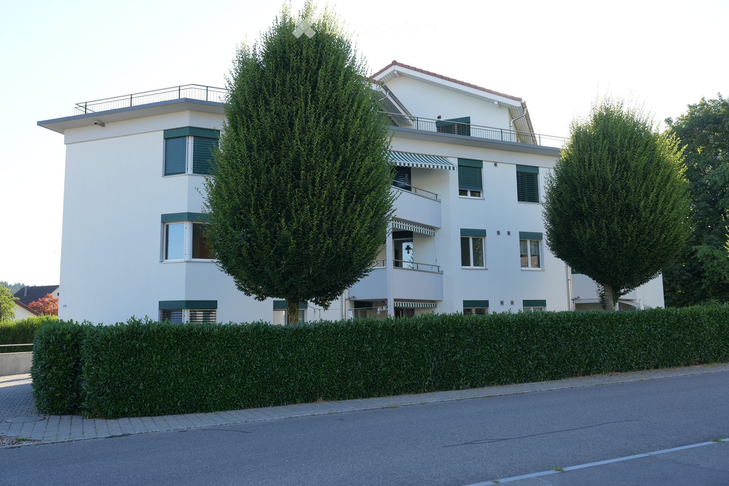 Modern two-story house with white exterior, green shutters, balconies, and surrounding greenery.