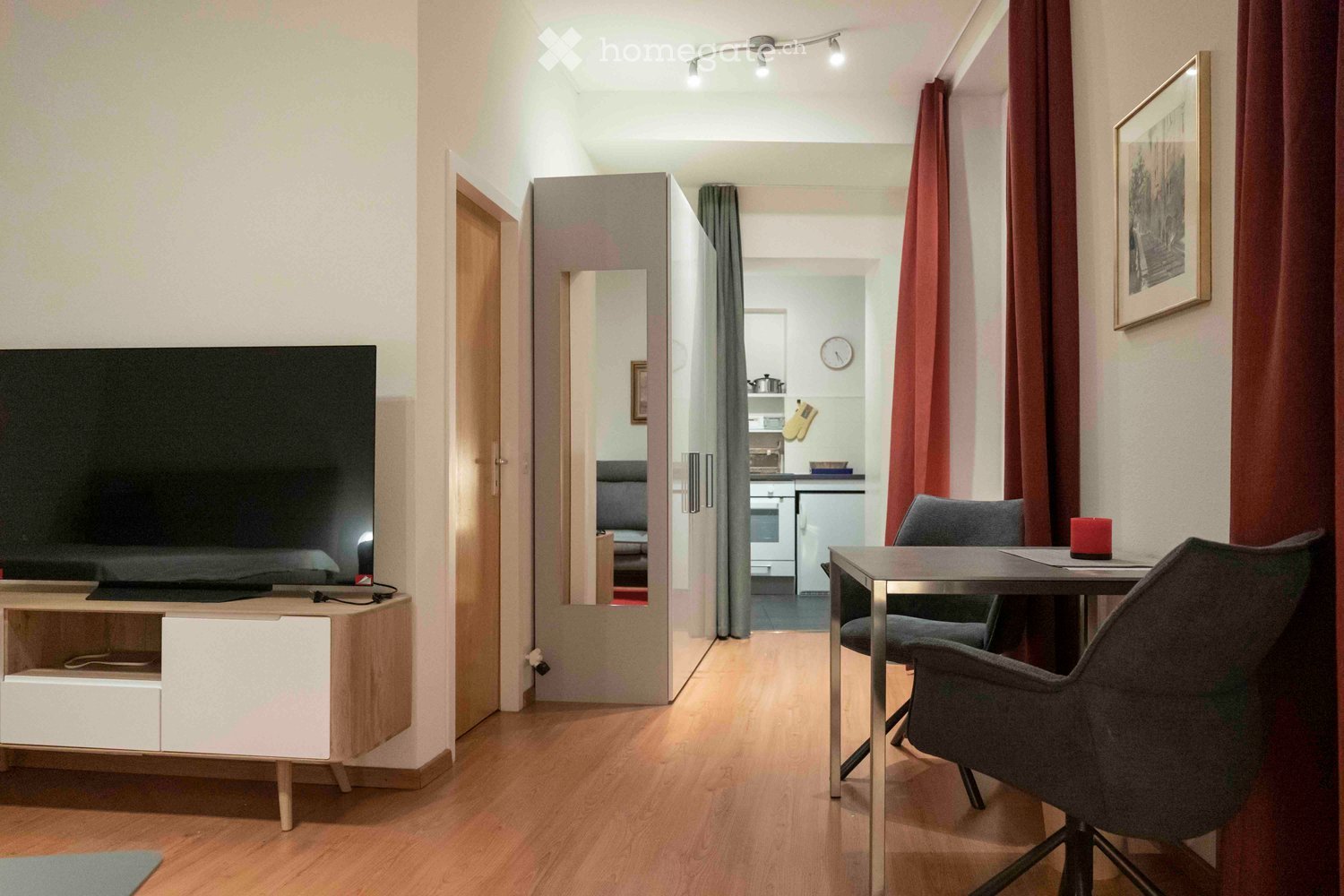A modern, furnished room with a large flat-screen TV on a wooden stand, a small dining area with a table and chairs, and an open kitchen area visible in the background. The room features wooden flooring, warm lighting, and red curtains.