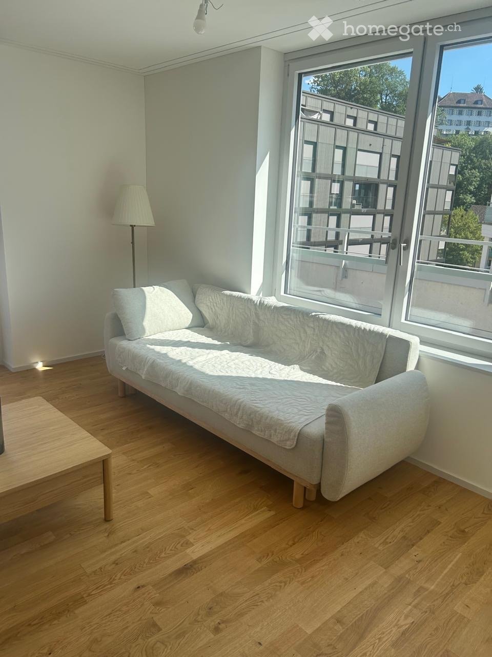 Bright living room with wooden flooring, a grey sofa with white cushions, a small wooden table, and a floor lamp near the window offering a view of the balcony and surrounding buildings.