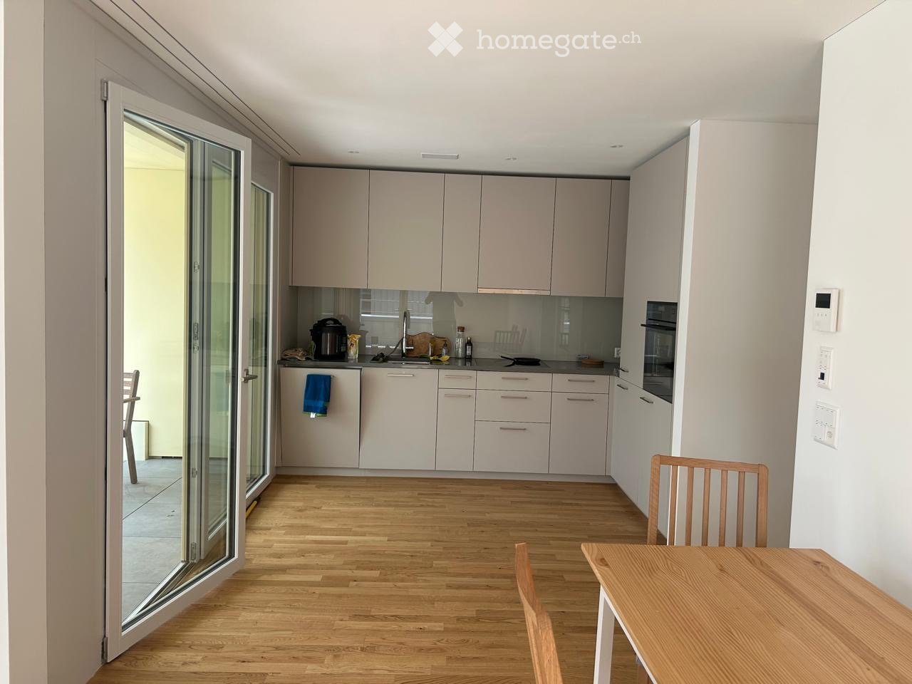 Modern kitchen with light-colored cabinets, a wooden dining table, and chairs, featuring a large sliding glass door leading to a balcony.