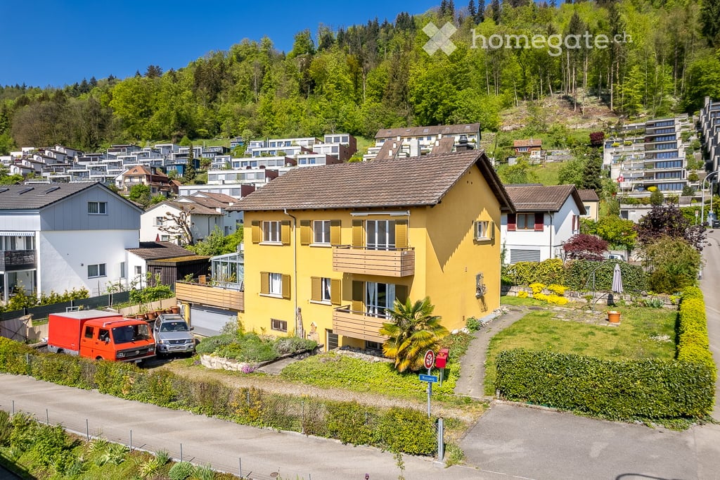 Bright yellow two-story house with a brown tiled roof, multiple windows, and a balcony. The house is surrounded by a well-maintained garden and is situated in a hilly, green neighborhood with other houses and trees in the background.