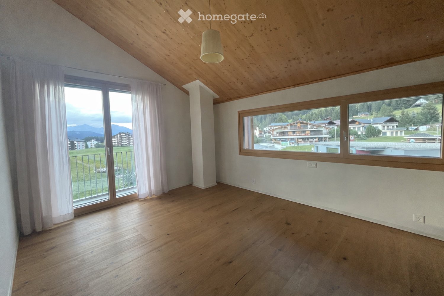 Empty room with wooden flooring, white walls, large windows, and a hanging light fixture.