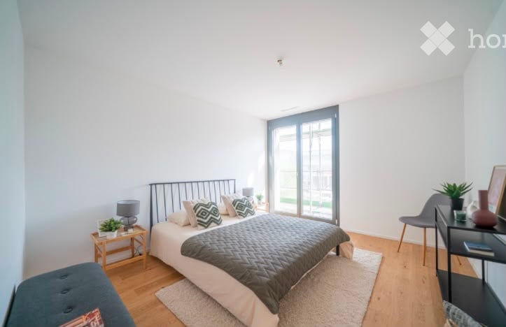 Bright, modern bedroom with a large bed, nightstand, lamp, chair, small table with plant, and picture frame on the wall. The room features wooden flooring and large windows with a view of the outdoor area.