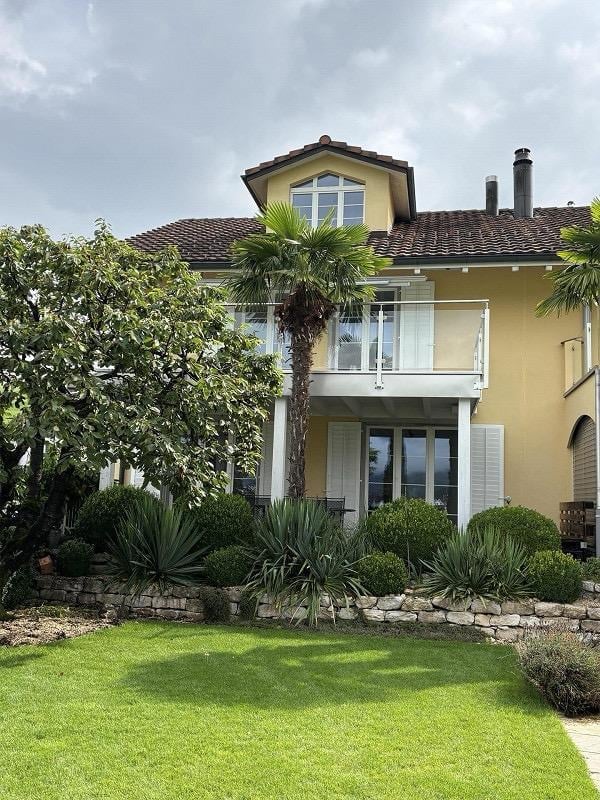 Two-story house with a tiled roof, yellow exterior, and a balcony on the upper level. The front yard features a well-maintained lawn, a stone wall, and various plants, including a palm tree.