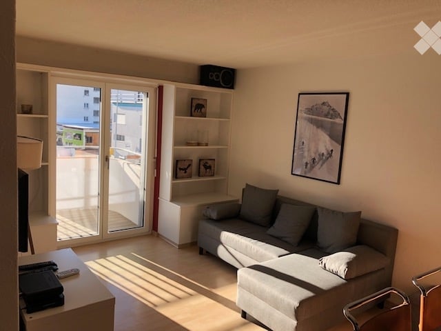 Sliding glass door leading to a balcony, white wall, gray couch, gray ottoman, white wooden table, shelf, painting, remote