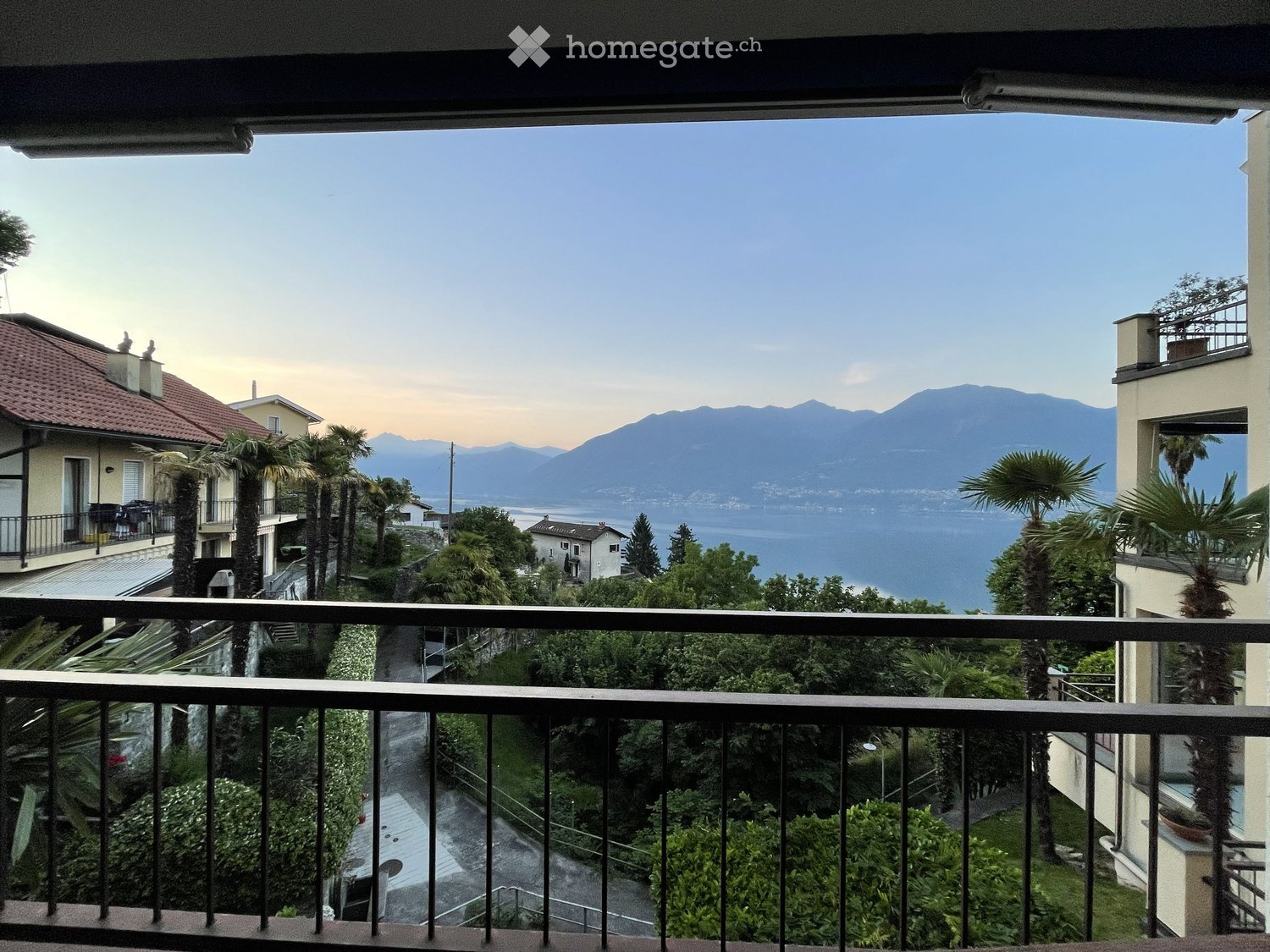 Balcony overlooking a scenic view of a lake, mountains, and surrounding buildings with palm trees and greenery.