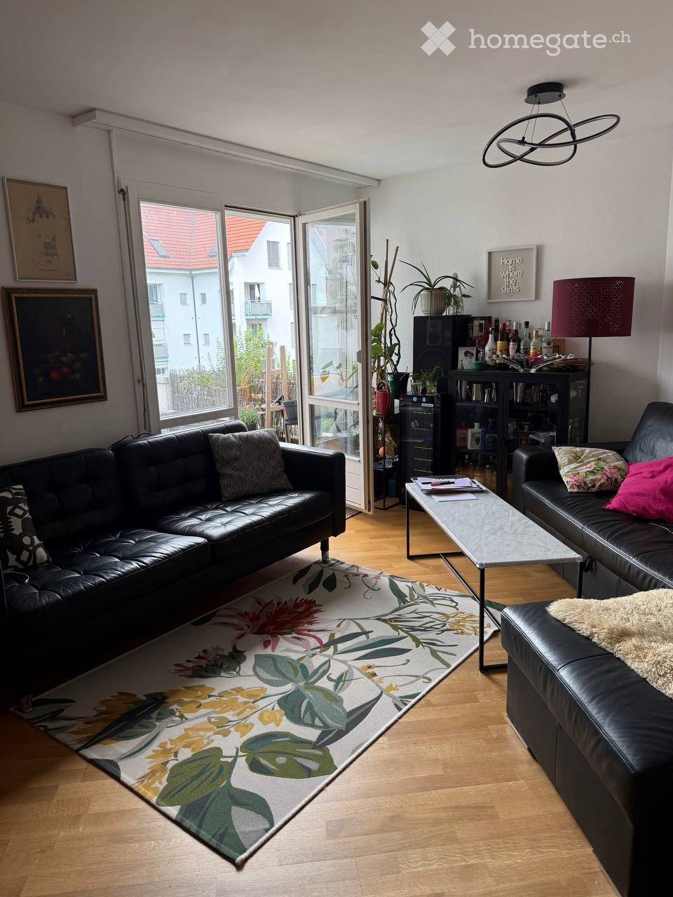 Modern living room with wooden flooring, large windows, black leather sofas, coffee table, floor lamp, bookshelf, and decorative plants.