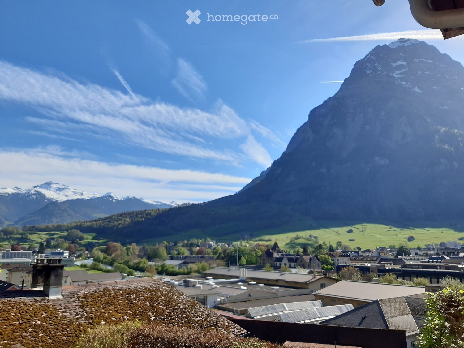 Panoramic view from a balcony showing a town with traditional houses, green fields, and snow-capped mountains in the background.