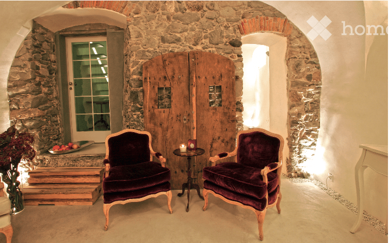 A cozy, rustic room with stone walls and arched doorways. Two plush armchairs in deep purple velvet are positioned facing each other, with a small round table between them holding a candle. A fruit bowl and vase with flowers sit on a wooden bench near the