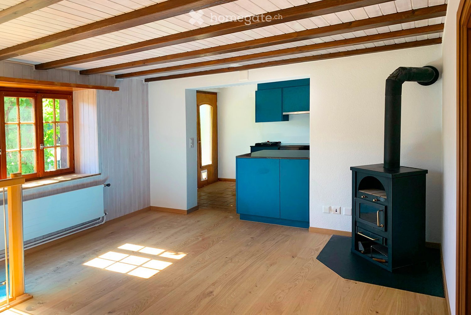 Bright room with wooden floors, exposed wooden beams, a blue cabinet, and a wood stove with a chimney. Large windows allow natural light to fill the space.