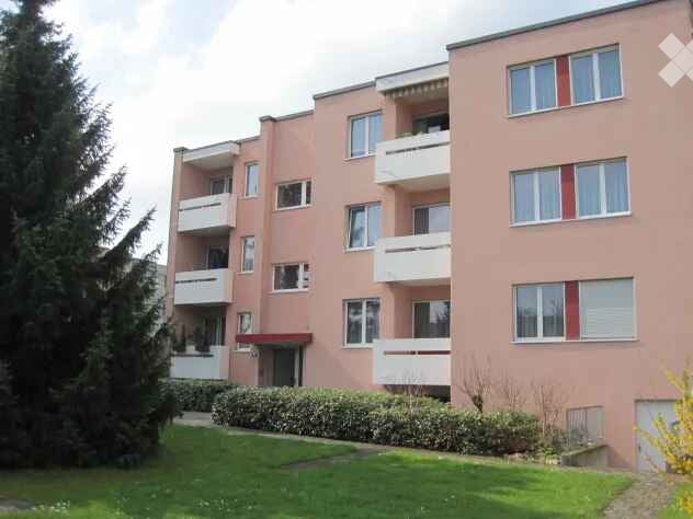 Pink apartment building with multiple balconies and windows, surrounded by greenery and a well-maintained lawn.
