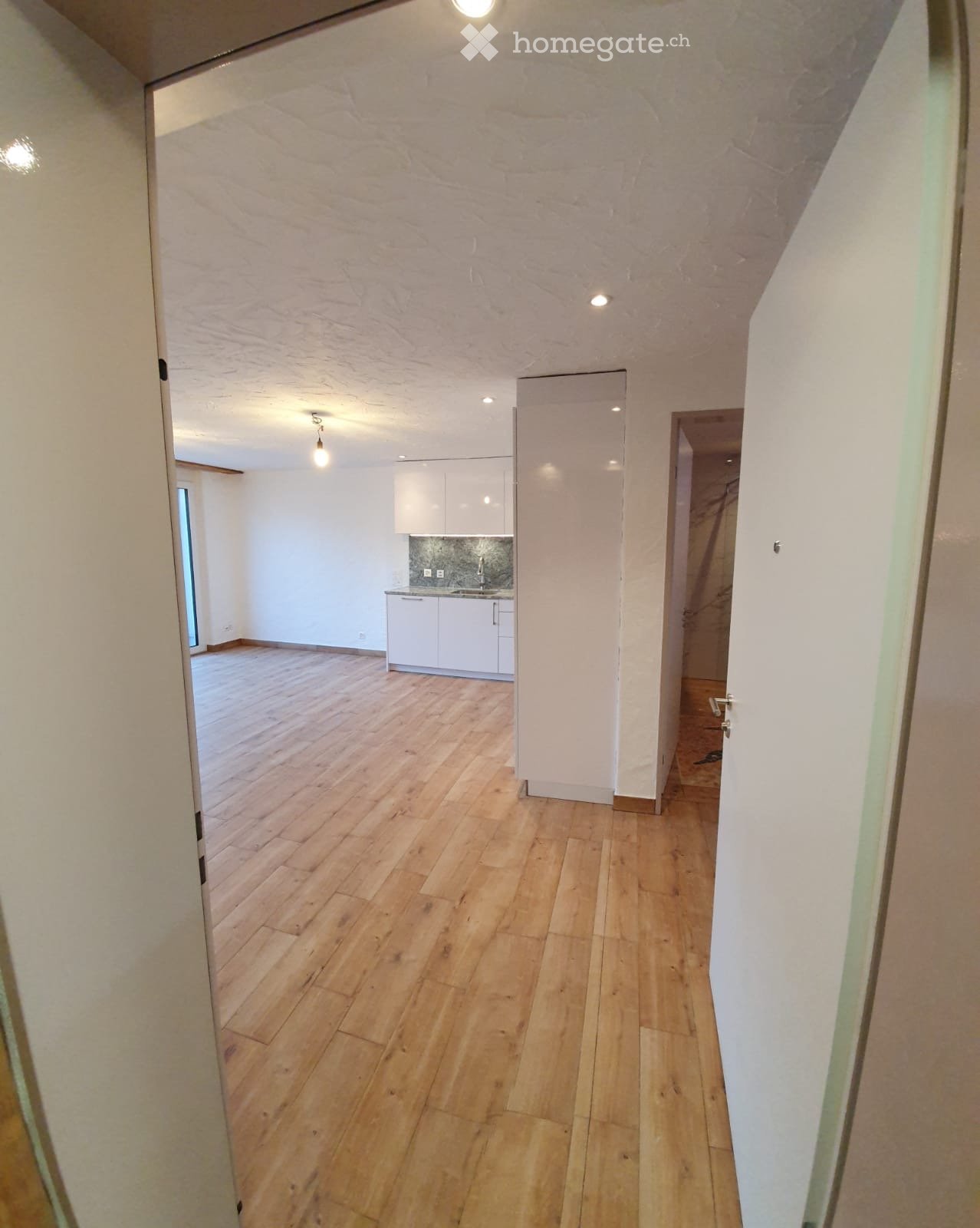 Empty hallway with wooden flooring and white walls, leading to a small kitchen area with white cabinets and a sink.