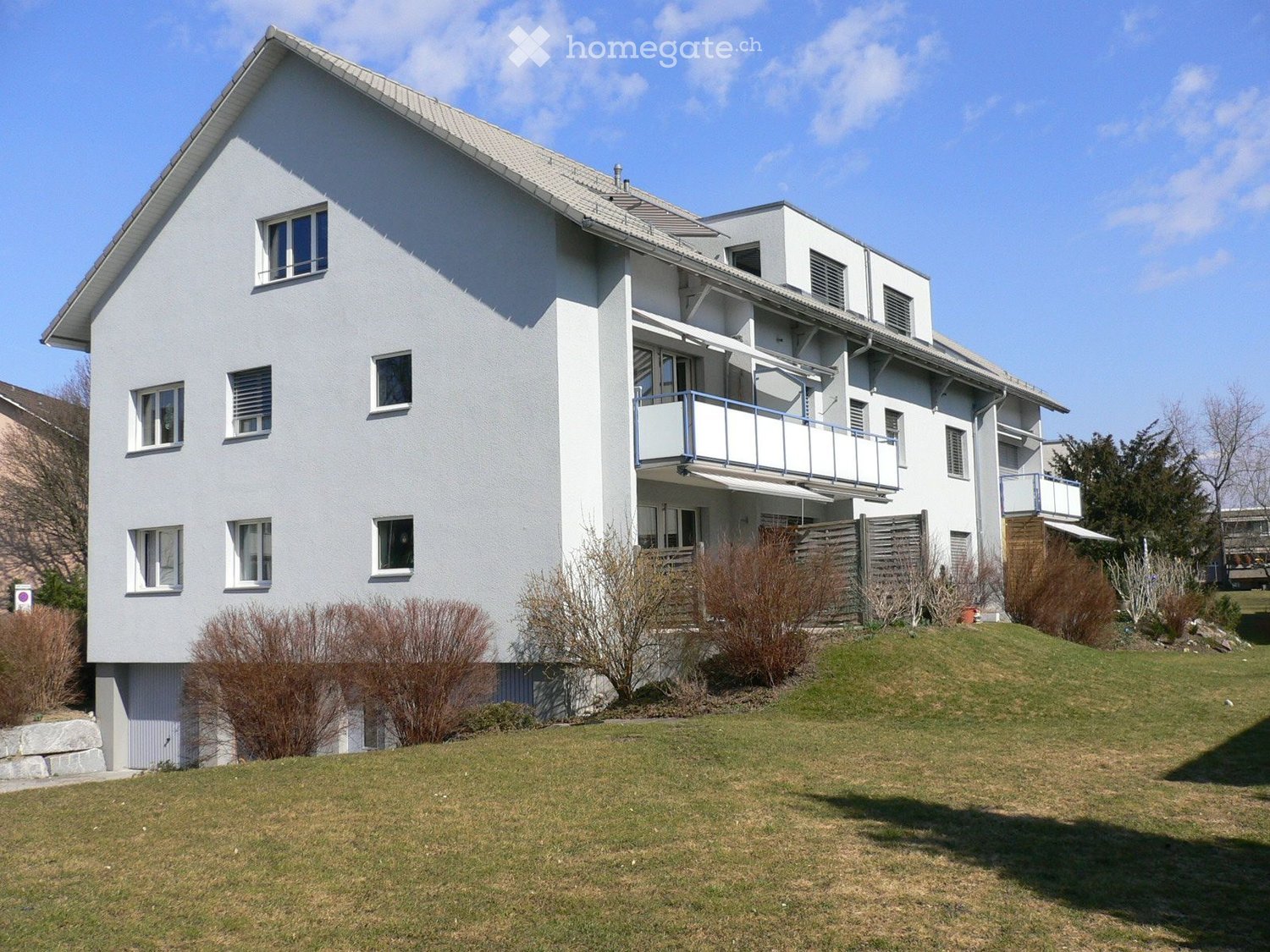 Two-story modern house with white exterior, multiple windows, balconies, and a well-maintained lawn.