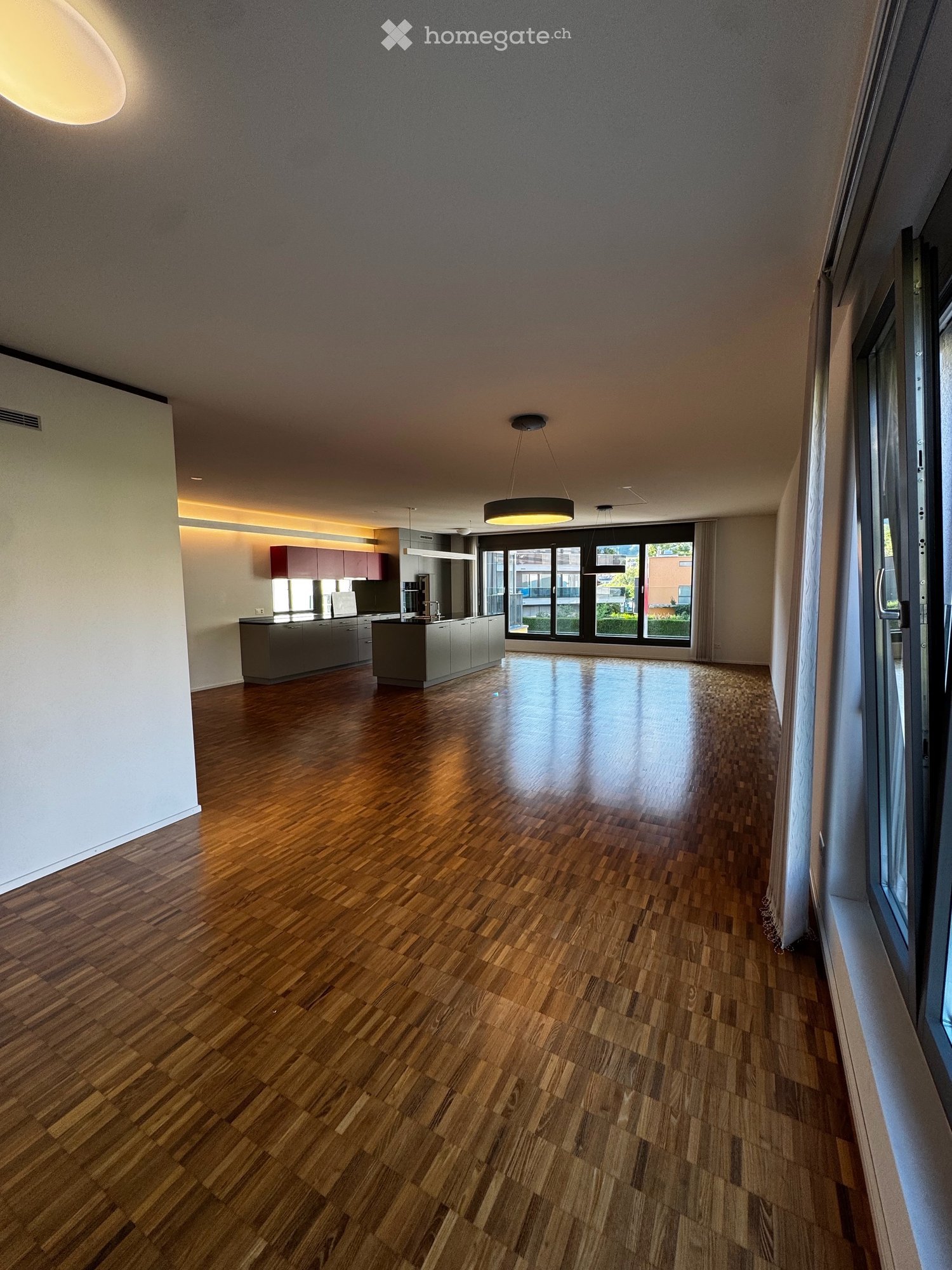 Open-plan space with wooden flooring, modern kitchen area with cabinets and appliances, large windows with sliding doors leading to a balcony, and recessed lighting.