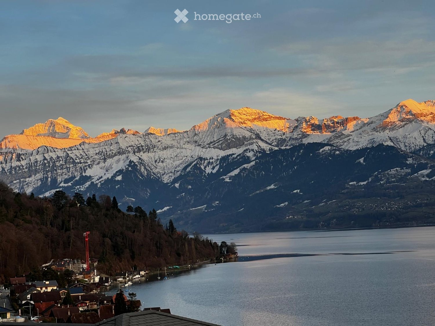 Panoramic view of snow-capped mountains and a calm lake, with a small village on the shore.