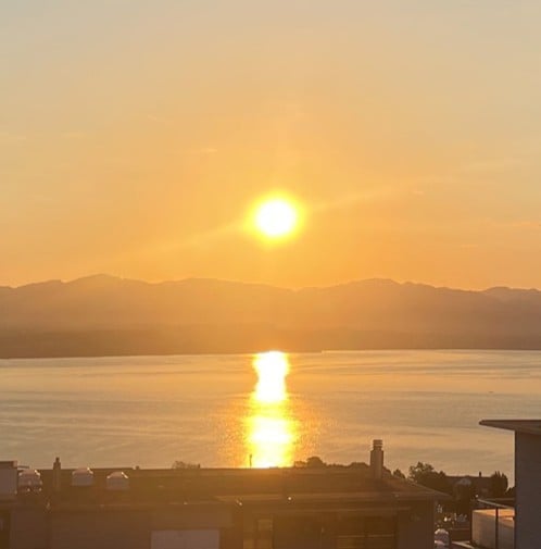 Balcony or terrace view overlooking a lake at sunset, with mountains in the background and the sun reflecting on the water.