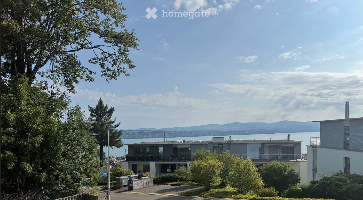 Modern two-story house with large windows and balconies, situated on a hillside overlooking a body of water and distant mountains.