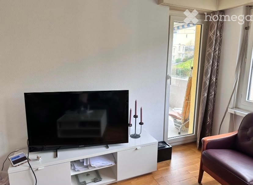A modern living room with a flat-screen TV on a white cabinet, a purple armchair, and a candle holder on the table. The room has wooden flooring and large windows with curtains.