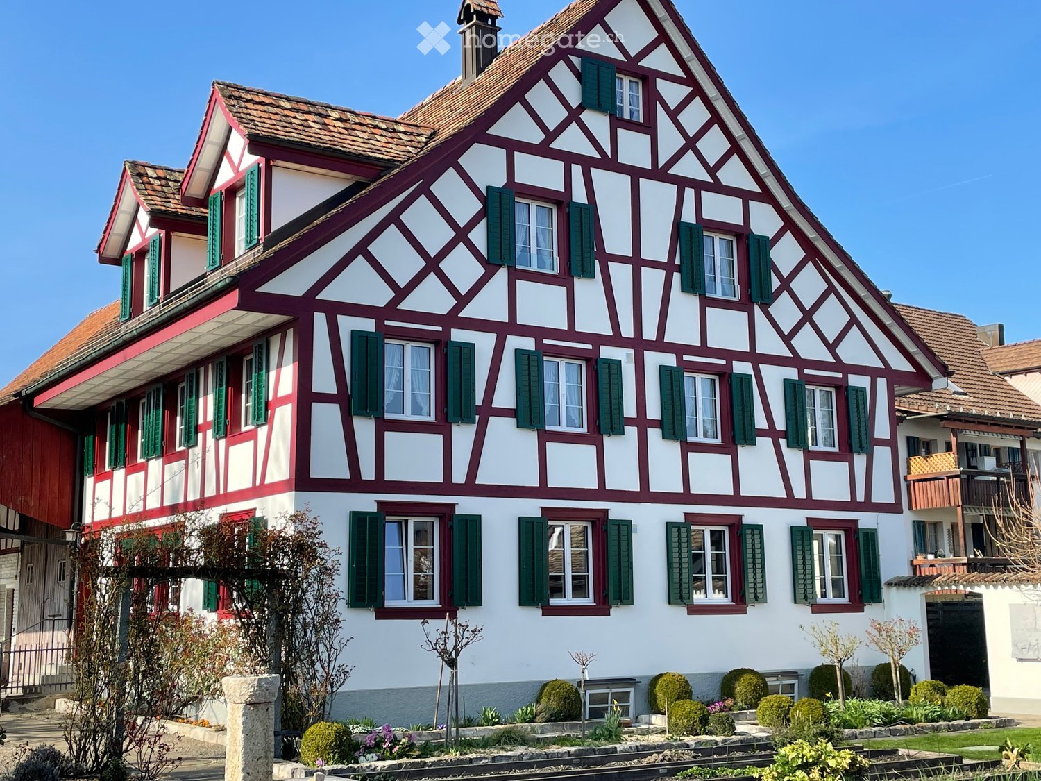 Traditional half-timbered house with red and white exterior, multiple windows with green shutters, and a well-maintained garden in front.