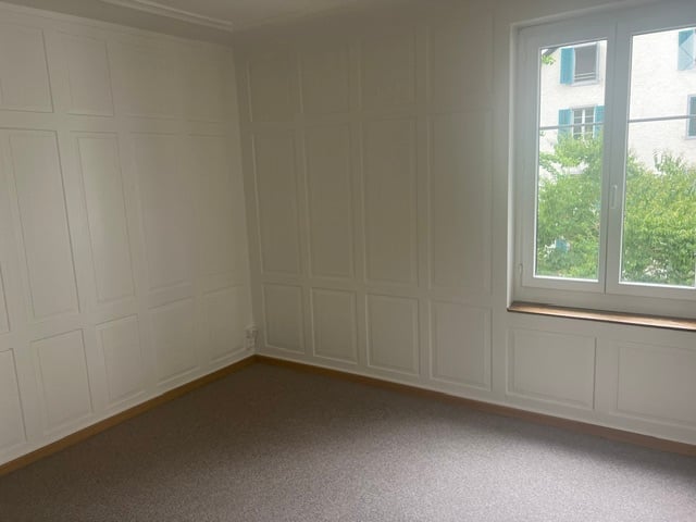 Empty room with white paneled walls, carpeted floor, and a large window with a view of greenery.
