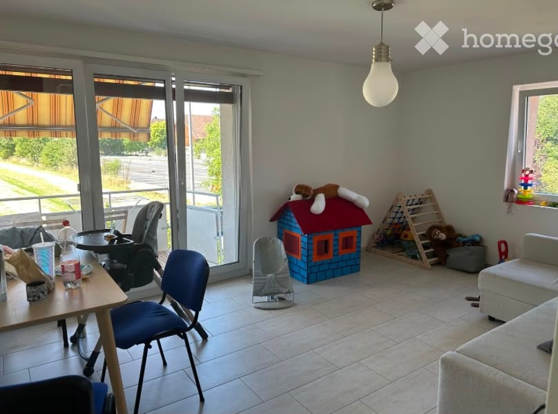 Bright living room with large sliding glass doors leading to a balcony, a sofa, a small table with chairs, a baby chair, a toy house, and various toys including a ladder and stuffed animals.