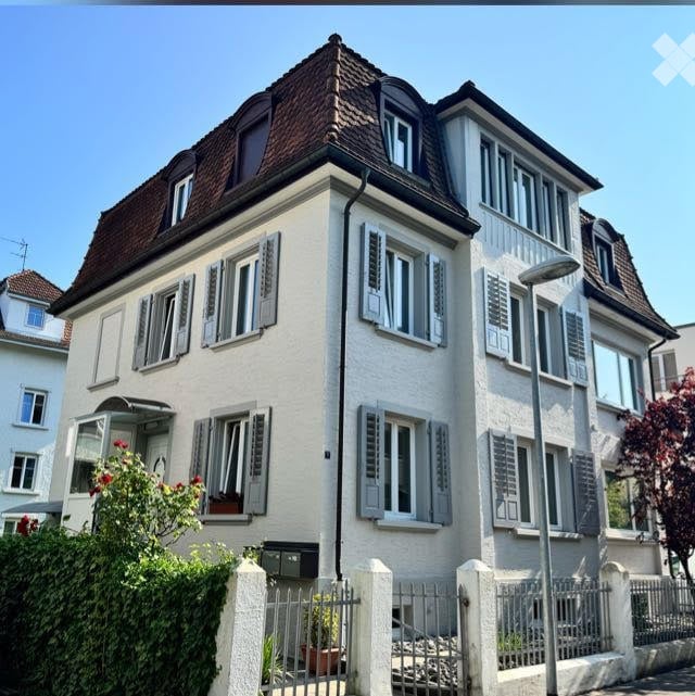 A multi-story residential house with a classic European design, featuring white walls, grey shutters, and a brown tiled roof. The house has multiple windows and a small garden area in front with a low fence.