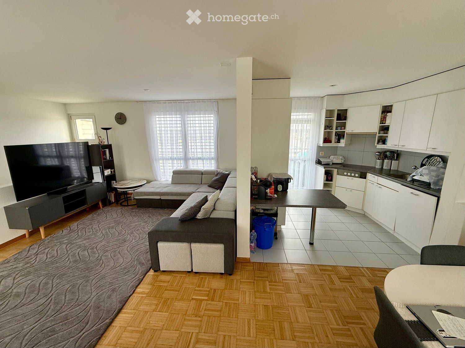 Open-plan living and dining area with a grey sectional sofa, coffee table, TV stand, and a small dining table with chairs. The kitchen features white cabinets, a stove, sink, and fridge.