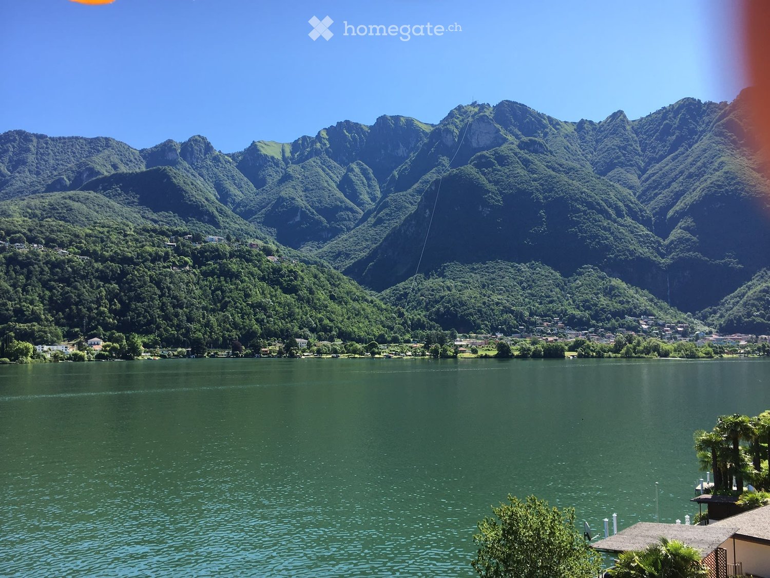A scenic view of a lake surrounded by lush green forests and rugged mountains under a clear blue sky.