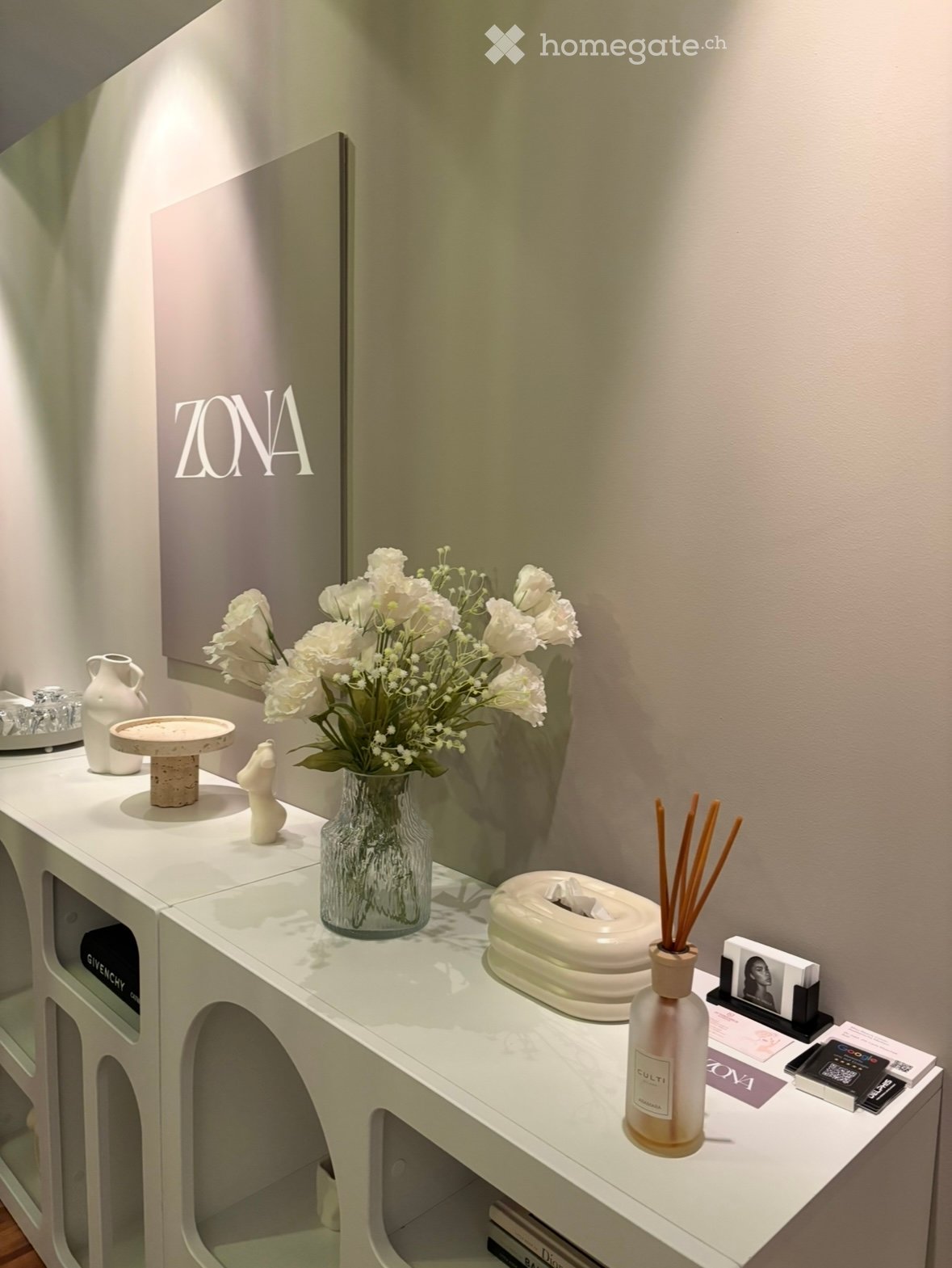 A modern, minimalist vanity area with a white table, a vase of white flowers, a decorative bottle, a mirror, and various small decorative items.