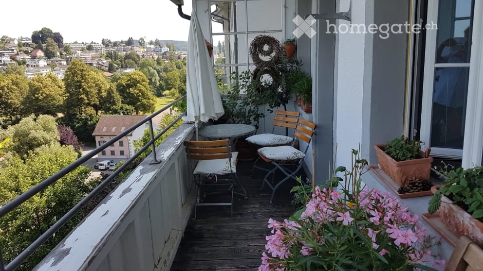 Balcony with wooden floor, metal railing, small round table, two wooden chairs, potted plants, and a view of the city and greenery.