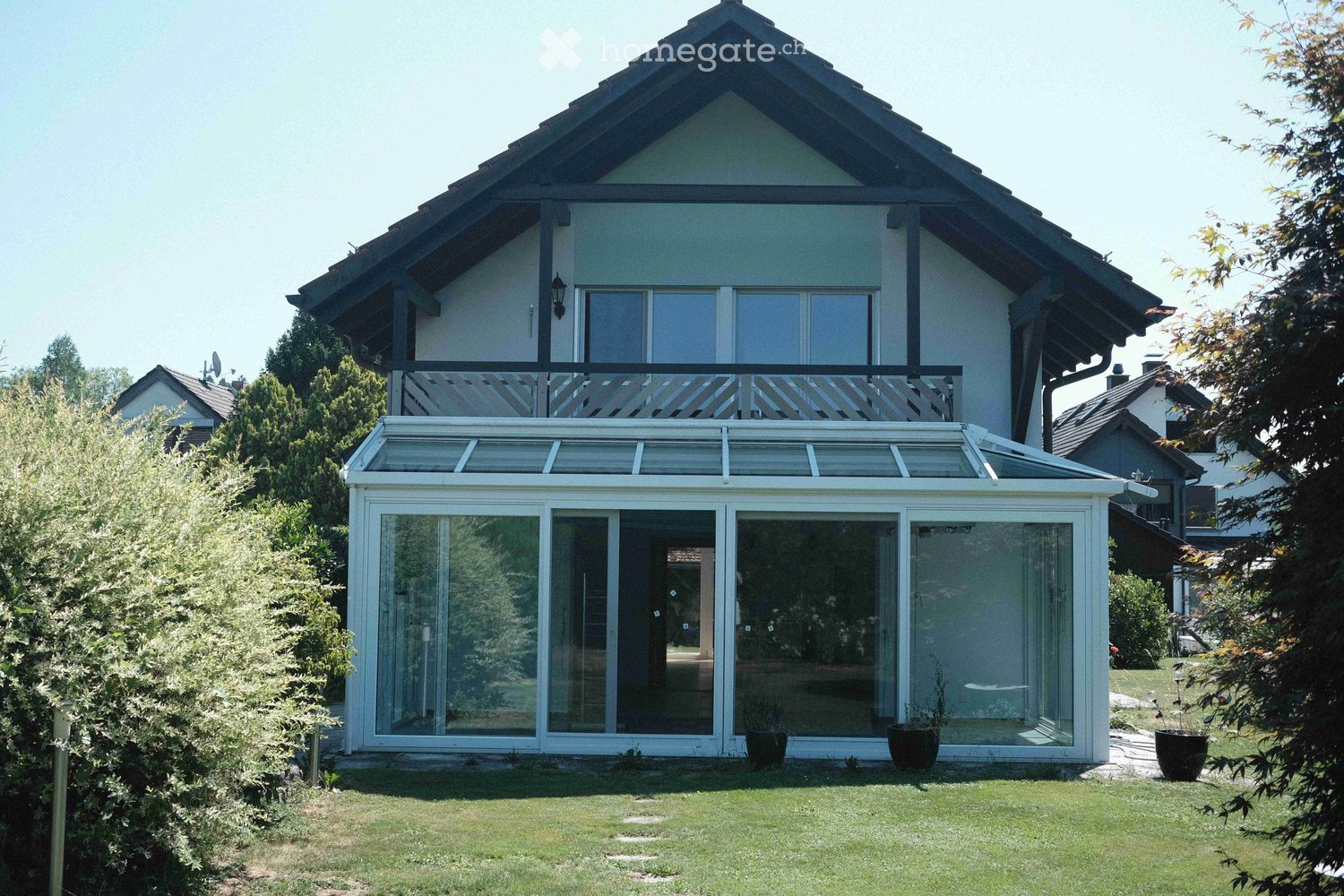 Two-story house with a balcony and a glass-enclosed patio area. The patio has a retractable roof and is surrounded by greenery.
