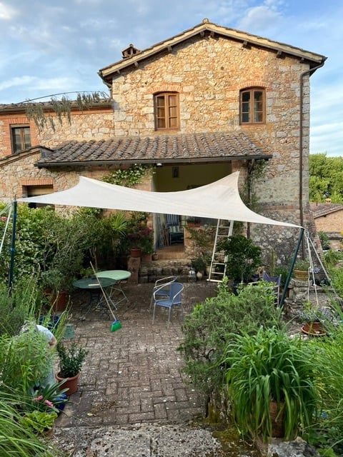 Outdoor patio area with a stone house in the background, featuring a large white canopy, several potted plants, a small table, and chairs.