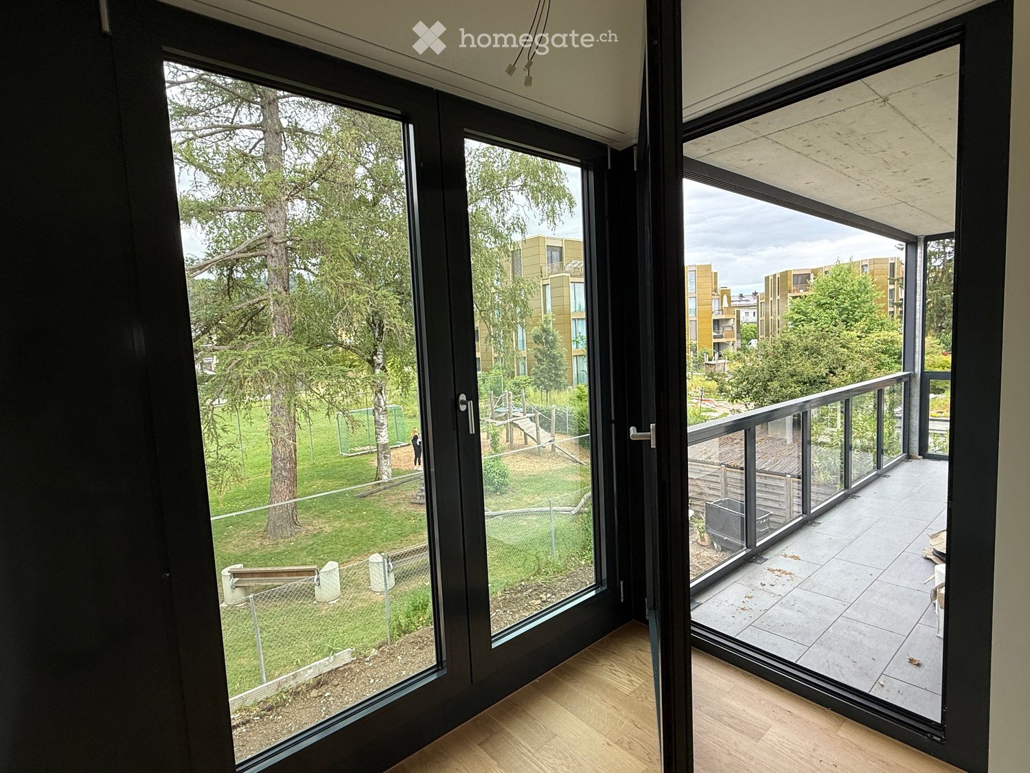 View from inside a room showing a modern balcony with large glass doors, wooden flooring, and a view of a green park area with trees and modern buildings.