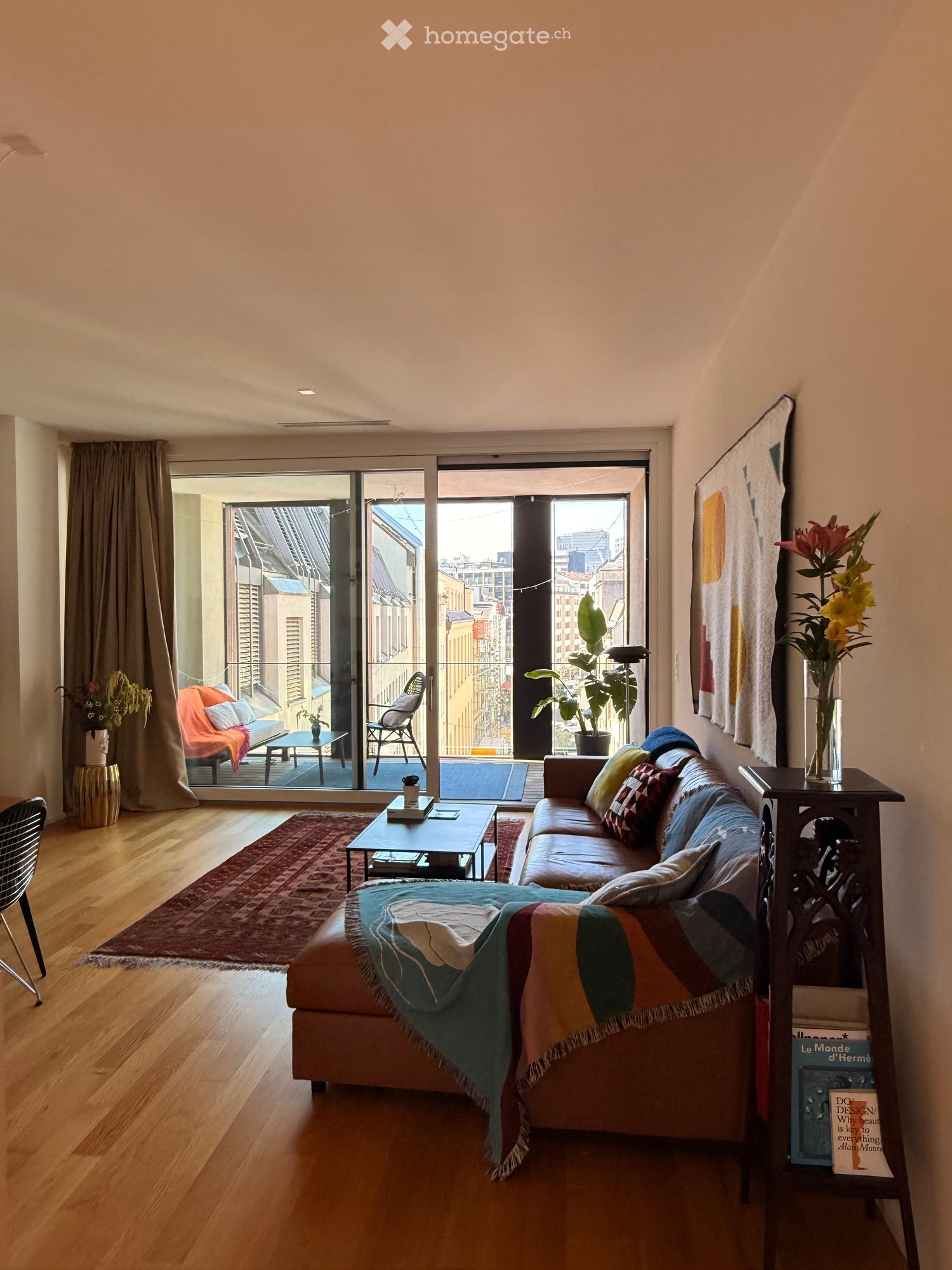 Modern living room with wooden flooring, large sliding glass doors leading to a balcony, a leather sofa with colorful cushions, a coffee table, and a side table with books and a vase of flowers.