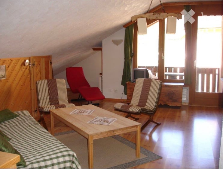 A cozy living room with wooden floors, a sofa, coffee table, chairs, and a television. The room features large windows with green curtains and a slanted ceiling typical of an attic space.